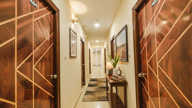 Um corredor de hotel com portas de madeira, um tapete xadrez, obras de arte emolduradas e uma pequena mesa com luminária e livros.