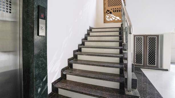 A staircase with a handrail, a wall-mounted elevator control panel, and a decorative panel with a lattice design. - Fabhotel Welcome Apartments (Mumbai)