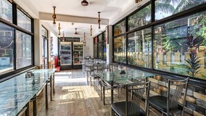 Un restaurante moderno con mesas y sillas de vidrio, amplias ventanas con vista a palmeras y un techo con lámparas colgantes.
