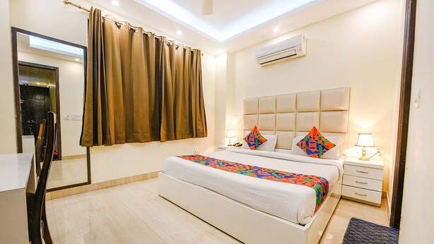 A hotel room with a large bed, a bedside table with a lamp, a nightstand, a mirror, and a window with curtains. - Fabhotel Ak Villa (New Delhi)