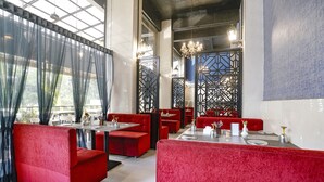 Ein Restaurant mit roten Polstersitzen, Tischen, großen Fenstern mit leichten Vorhängen und dekorativer Beleuchtung.
