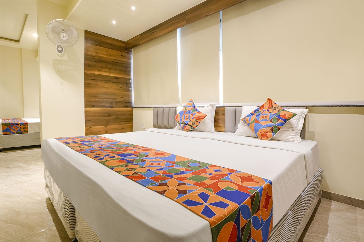 A neatly made bed with a colorful geometric-patterned bedspread, a wooden headboard, and a large window with blinds.