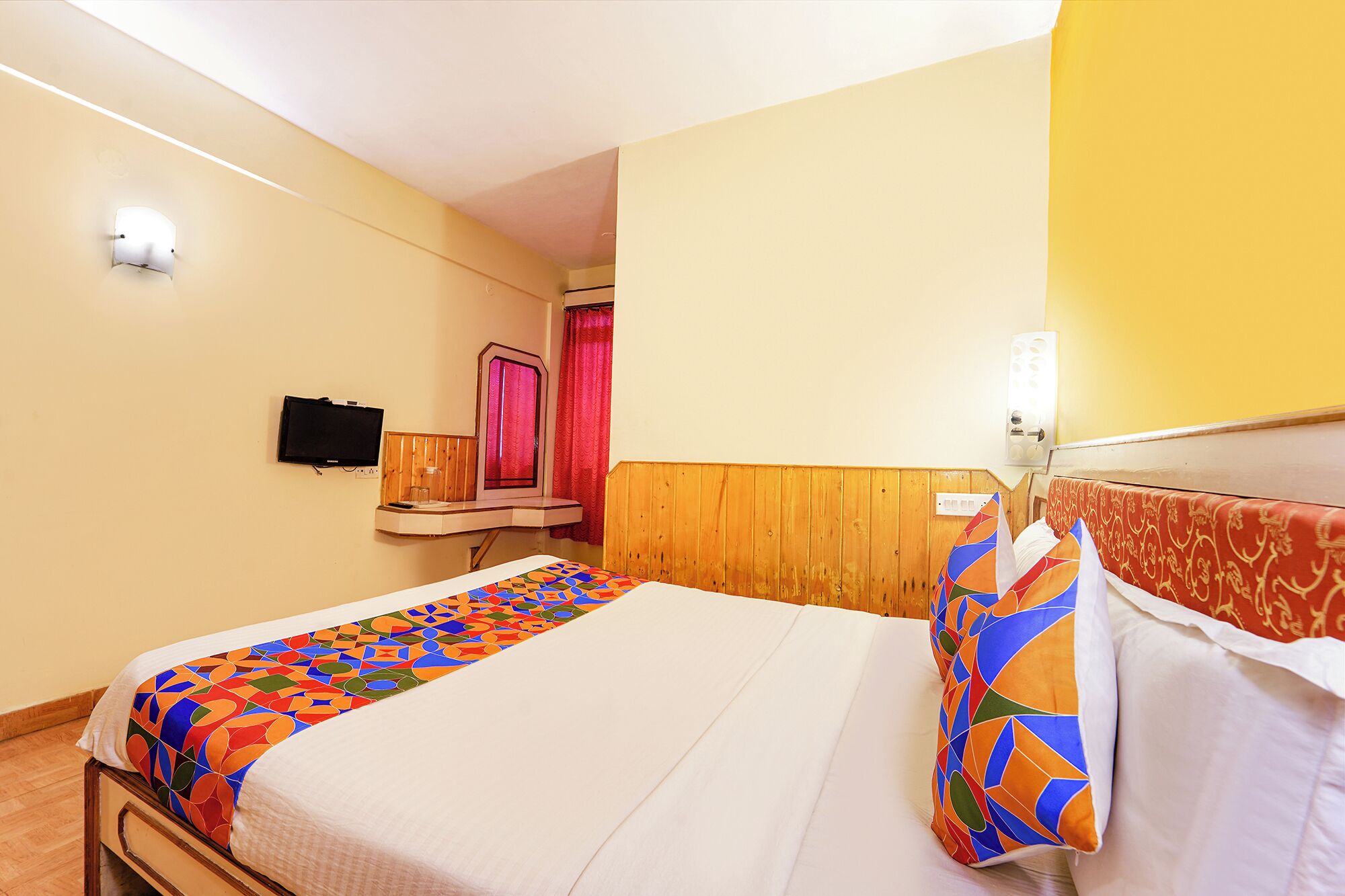 A hotel room with a bed, a bedside table, a mirror, a TV, and a window with curtains.