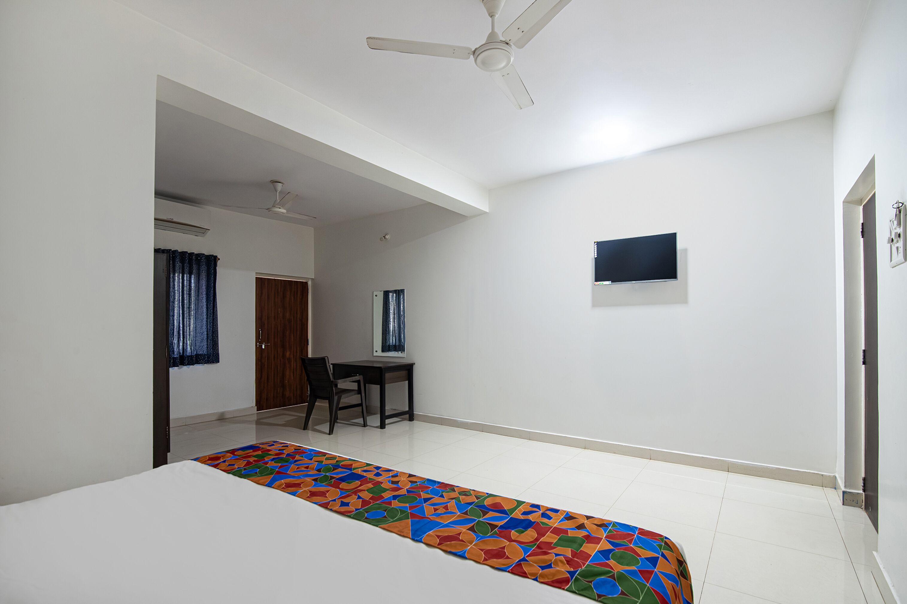 A room with a bed, a desk, a chair, a ceiling fan, and a TV mounted on the wall.