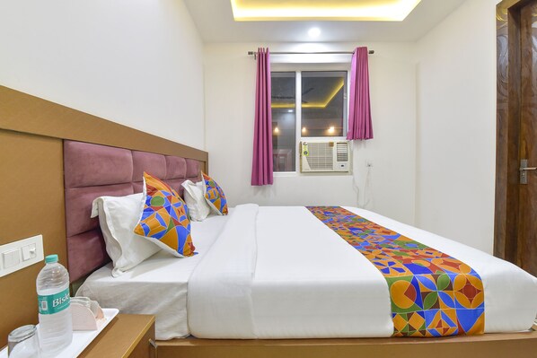 A hotel room with a large bed, a headboard, a window with curtains, an air conditioning unit, and a bedside table with a bottle and glasses. - Fabhotel Hari Residency (Haridwar)