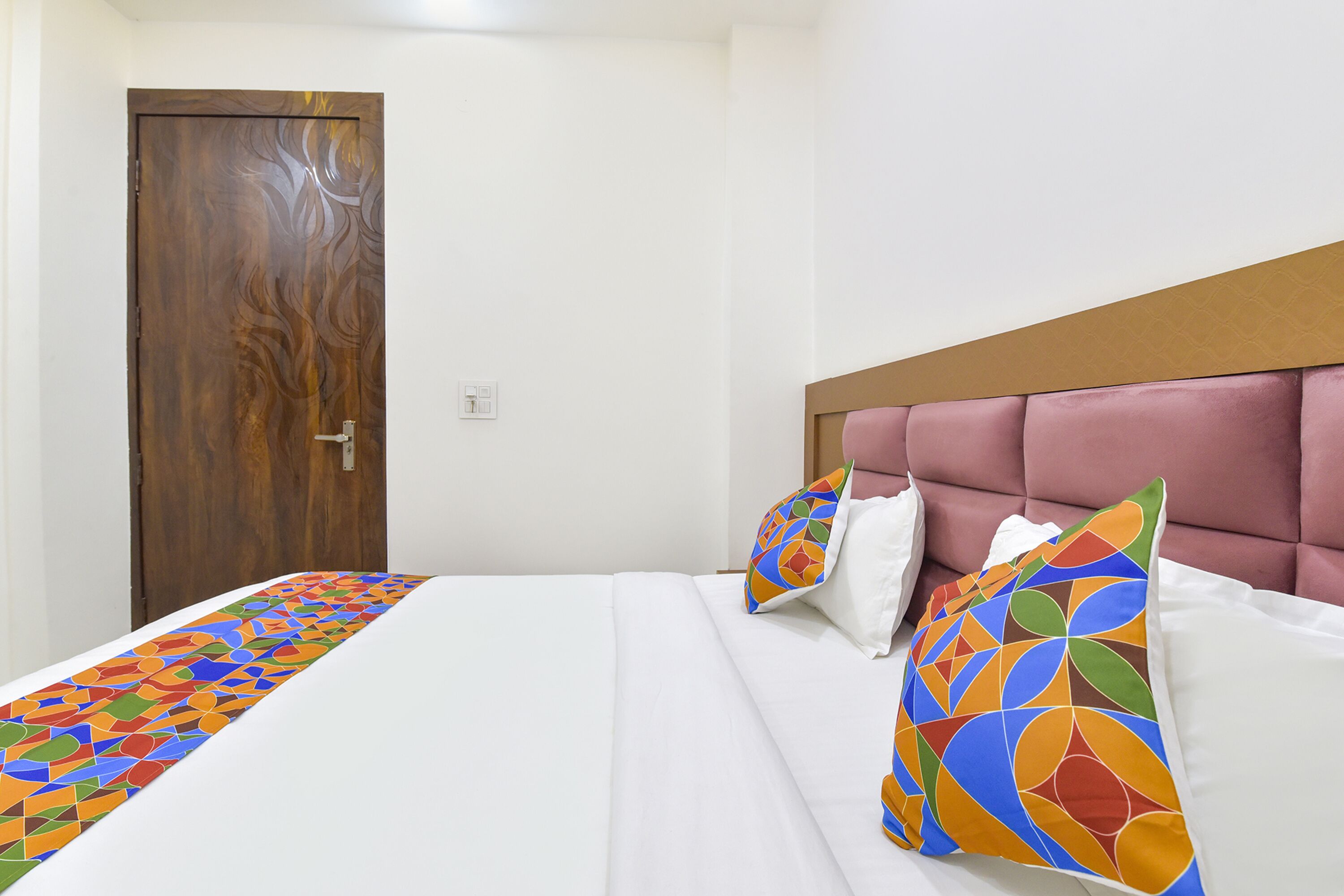A hotel room with a wooden door, a bed with white bedding, and colorful geometric-patterned pillows.