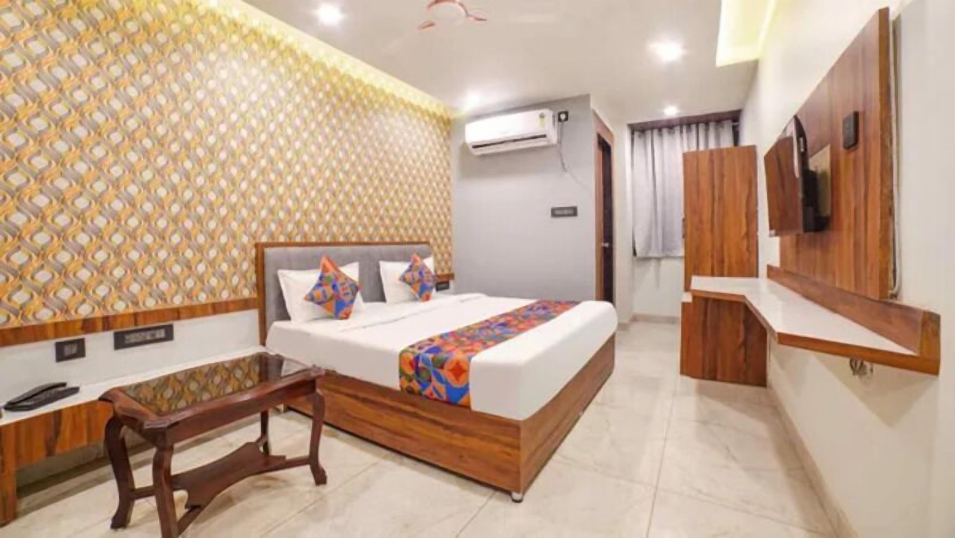 A hotel room with a bed, a wooden nightstand, a TV, and an air conditioning unit.