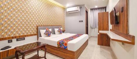 A hotel room with a bed, a wooden nightstand, a TV, and an air conditioning unit.