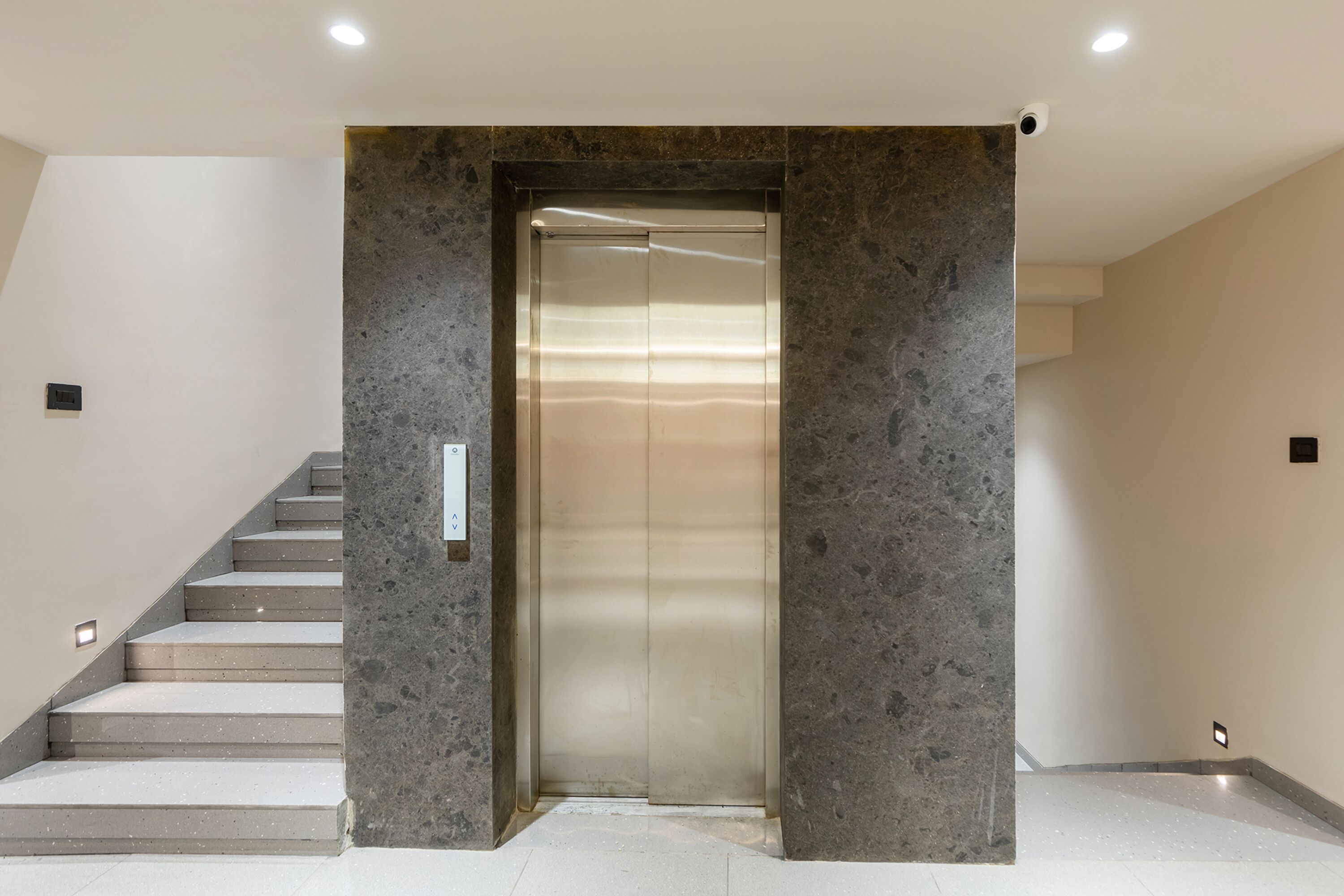 Ein moderner Aufzug mit metallischer Oberfläche, begleitet von einer Steinwand und einer Treppe.
