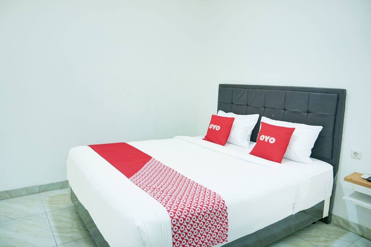 A neatly made bed with white and red bedding, including pillows with the word "OYO" on them, in a simple room with a plain wall and a small wooden nightstand.