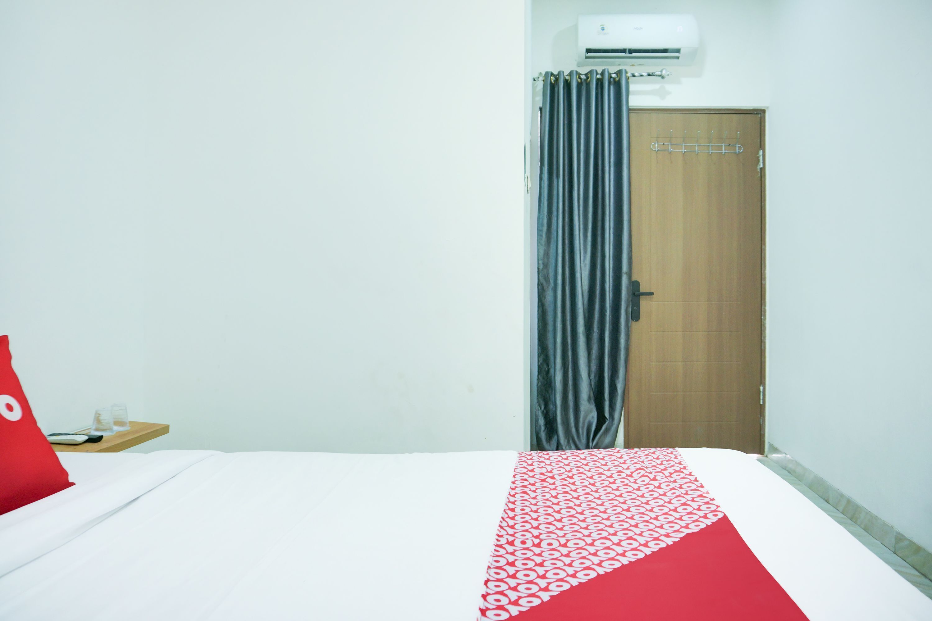 A hotel room with a bed, a red pillow, a wooden nightstand, a glass of water, and a door with a curtain.