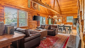 A log cabin interior with a wooden ceiling, leather sofas, a dining table, and a television.