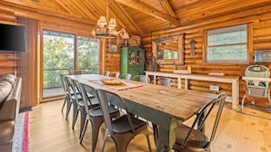 A wooden cabin interior with a dining table, chairs, a television, and a large window offering a view of trees.