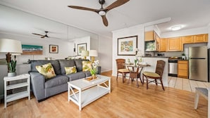 A modern living room with a grey sofa, white coffee table, and wooden dining table with chairs. There is a kitchen area with wooden cabinets and a stainless steel refrigerator.