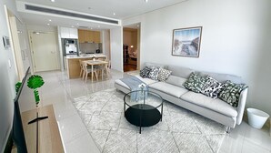 Een moderne woonkamer met een bank, een glazen salontafel en een eethoek, met een keuken op de achtergrond.