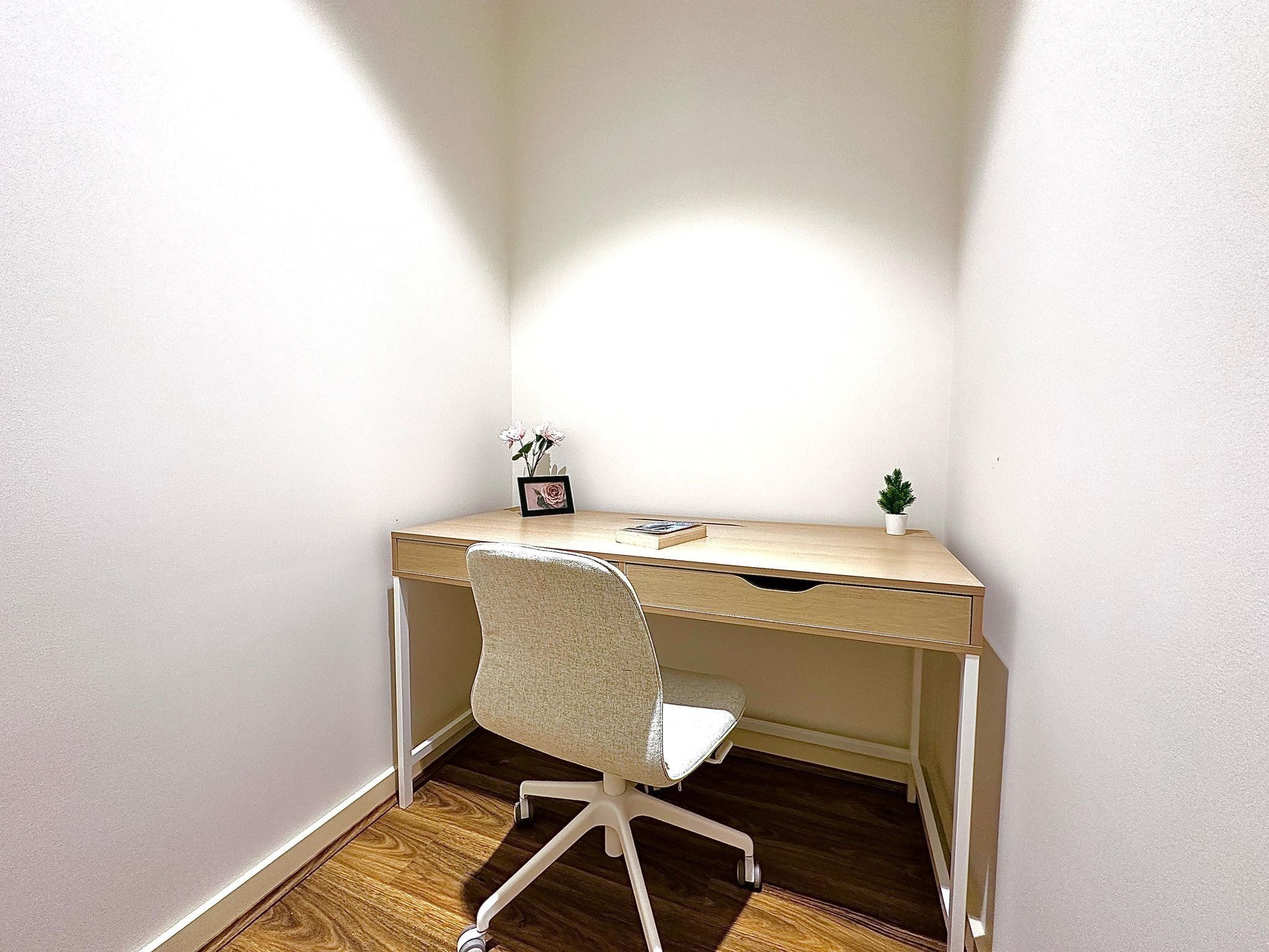 A corner office with a wooden desk, a white office chair, a small potted plant, and a framed picture on the desk.