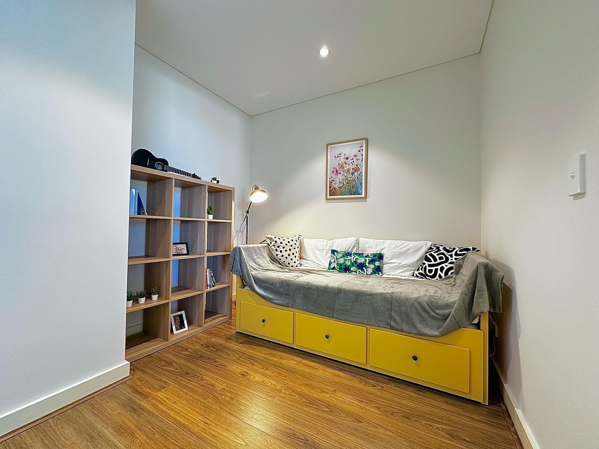 Une pièce avec un plancher en bois, un canapé jaune orné de coussins à motifs, une bibliothèque et un tableau encadré accroché au mur.