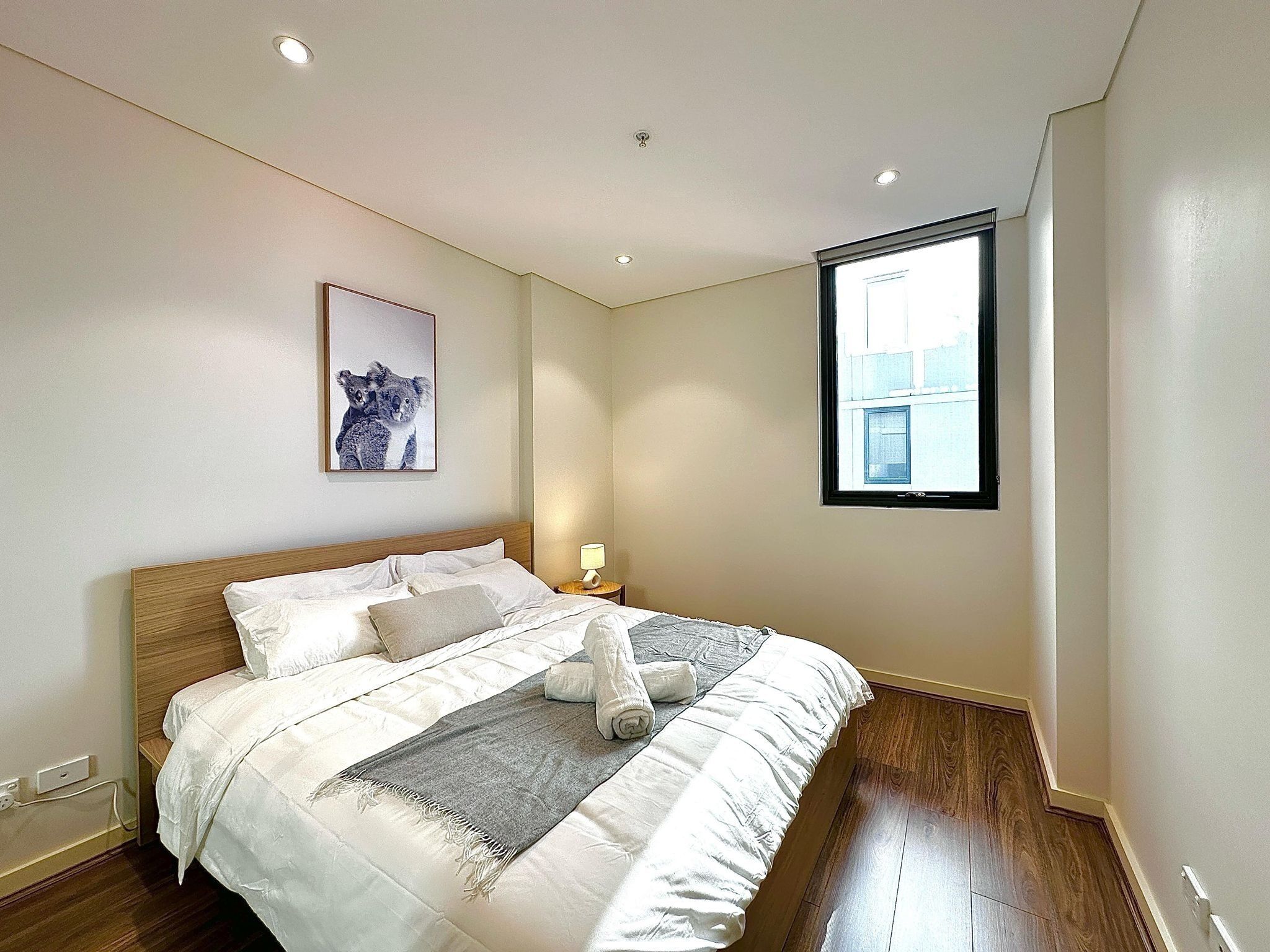 A neatly made bed with a grey throw blanket, a framed picture of koalas, and a window with a view of buildings.