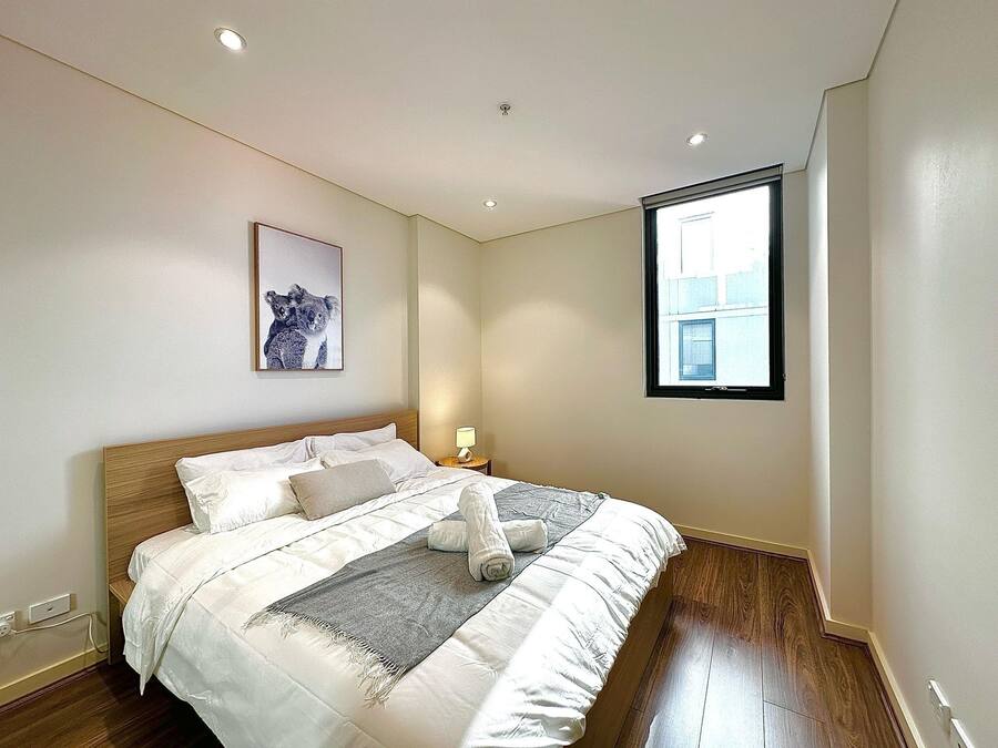A neatly made bed with a grey throw blanket, a framed picture of koalas, and a window with a view of buildings.