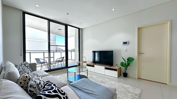 Een moderne woonkamer met een grote televisie, een bank met opvallende kussens, een glazen salontafel en een balkon met tuinmeubilair.