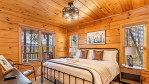 A wooden cabin bedroom with a metal bed frame, two bedside tables with lamps, a framed picture on the wall, and large windows with blinds. - Mountain Views, Dog friendly, Fenced Yard, Barrel Sauna, Hot Tub, Fire Pit
 (Ellijay)