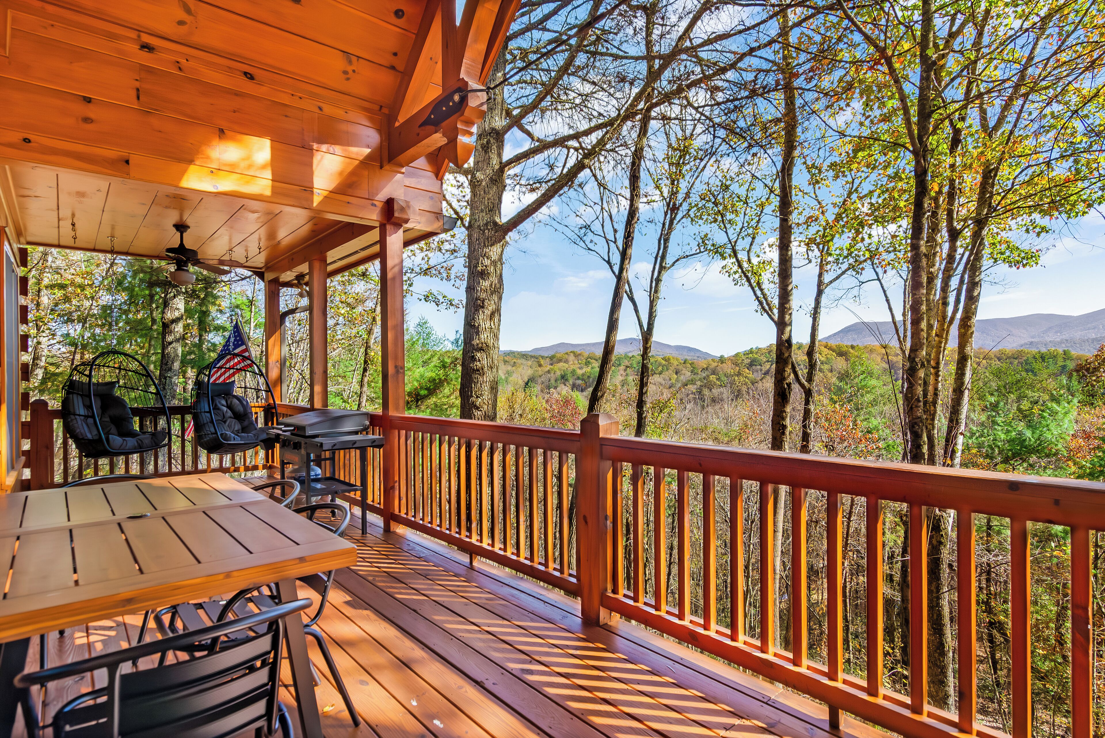 A wooden deck with a table and chairs, overlooking a forested area with trees and distant mountains.