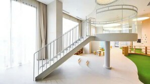A spacious indoor area with a central staircase, a green carpeted floor, and a glass ceiling structure. - The Antonia Apartments - Korea Town D7 (Ho Chi Minh City)