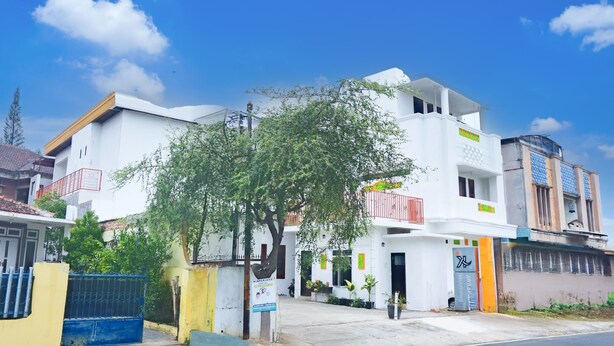 A white multi-story residential building with balconies, a blue gate, and a tree in the foreground.
