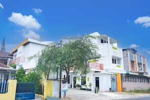 Un edificio residencial blanco de varios pisos con balcones, un portón azul y un árbol en primer plano.