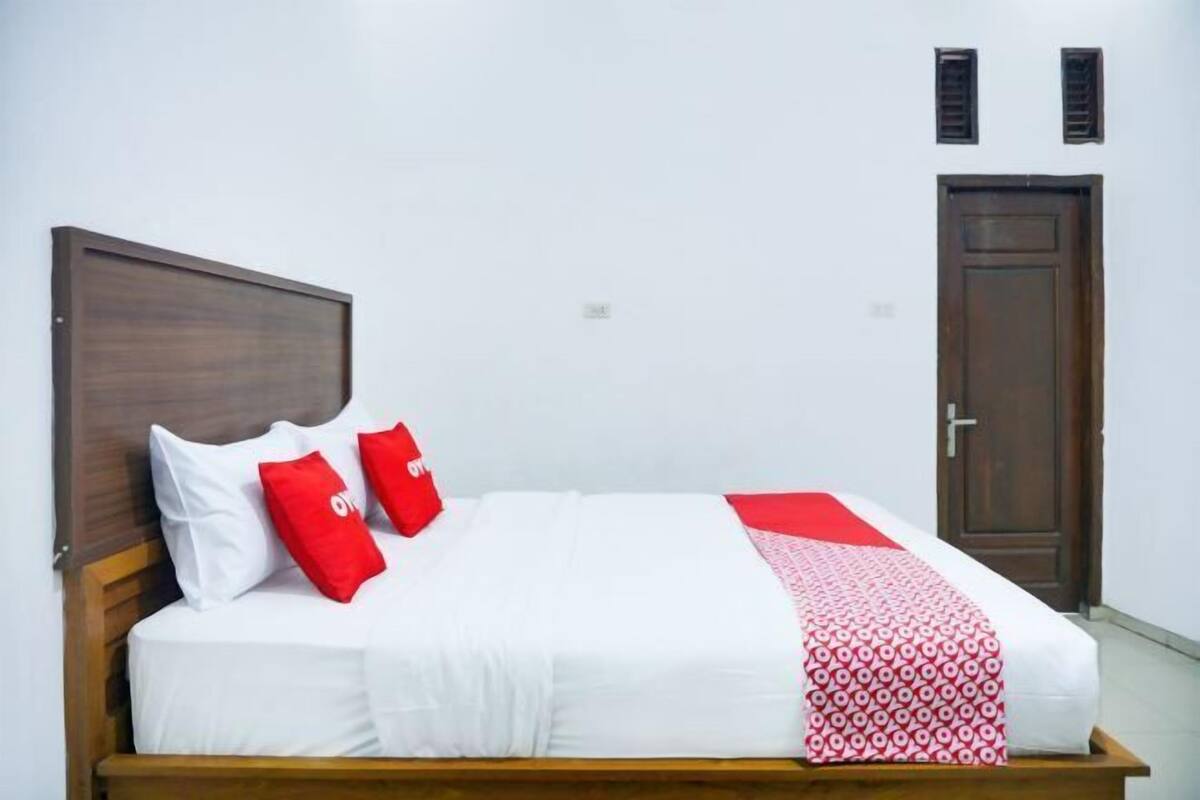 Uma cama com roupa de cama branca e detalhes vermelhos, uma cabeceira de madeira e uma porta com puxador.