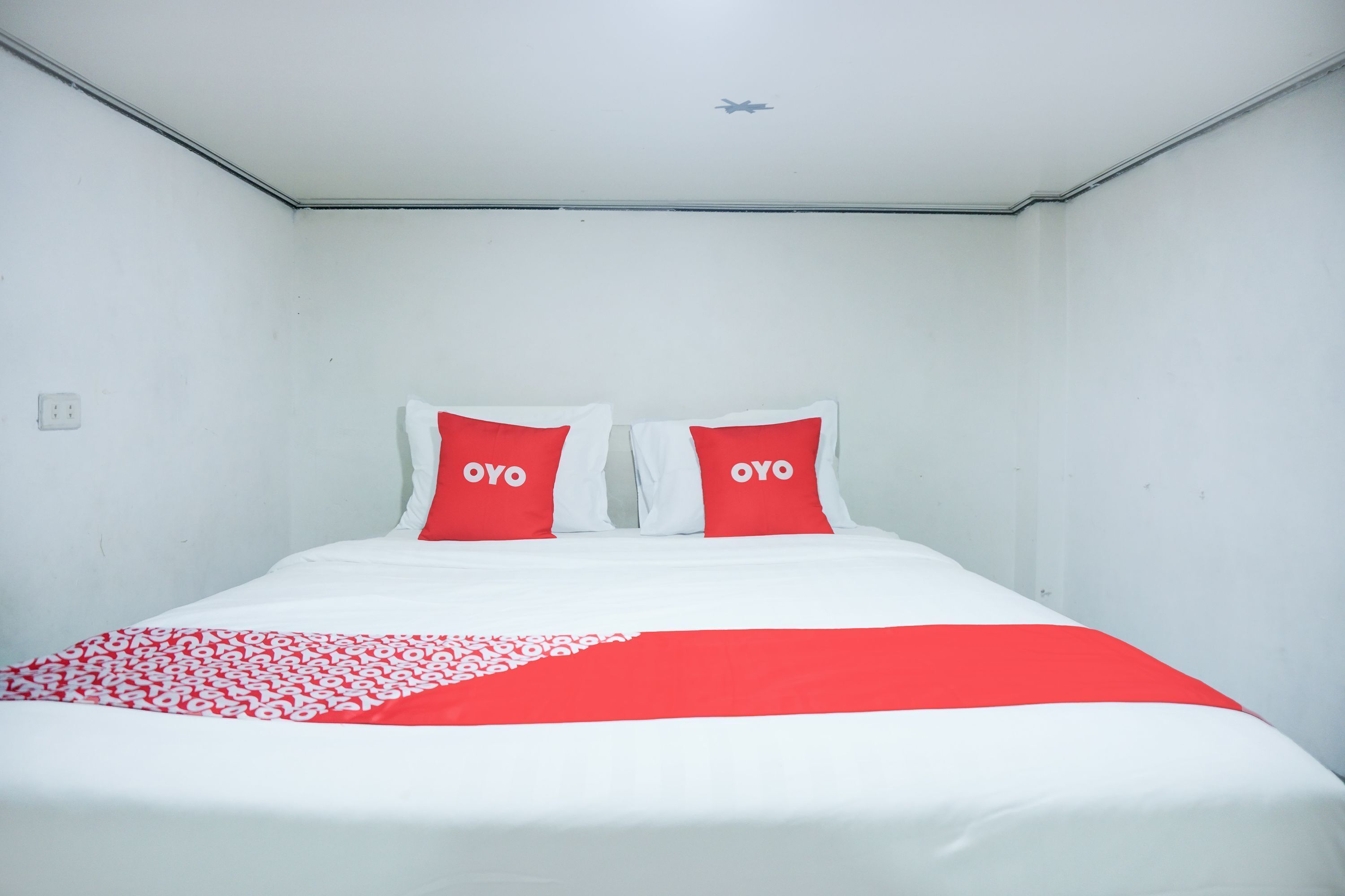 Habitación de hotel con cama, dos almohadas rojas con el texto "OYO", una colcha blanca y roja y un techo blanco con un adhesivo pequeño de un avión.