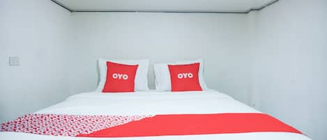 Habitación de hotel con cama, dos almohadas rojas con el texto "OYO", una colcha blanca y roja y un techo blanco con un adhesivo pequeño de un avión.