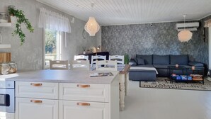 Una sala moderna con un sofá gris, una mesa de comedor con sillas, una zona de cocina con armarios y un gran ventanal que proporciona luz natural.