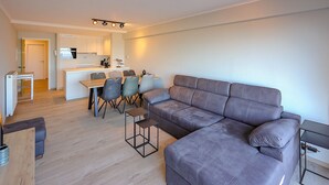 Een moderne woonkamer met een grijze modulaire bank, een eethoek met tafel en stoelen, en een keuken op de achtergrond.