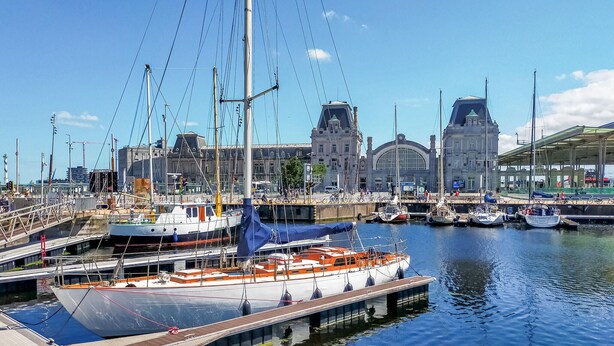Een jachthaven met aangemeerde boten, een brug en historische gebouwen op de achtergrond.