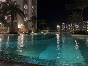 Vista noturna de uma piscina com água cristalina, rodeada por palmeiras e um edifício com janelas iluminadas.
