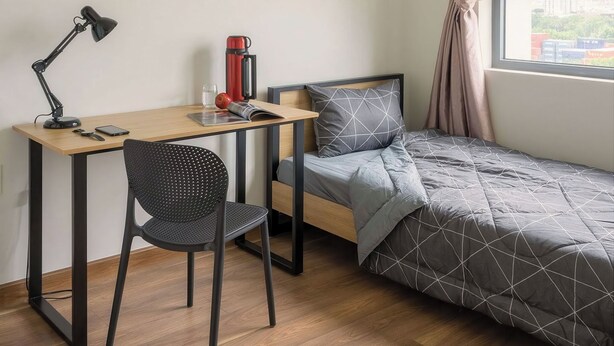 A compact room with a wooden desk, a black chair, a bed with a geometric patterned duvet, a red mug, a framed picture on the wall, and a window with curtains. - Saigon Riverside Complex D7 (Ho Chi Minh City)