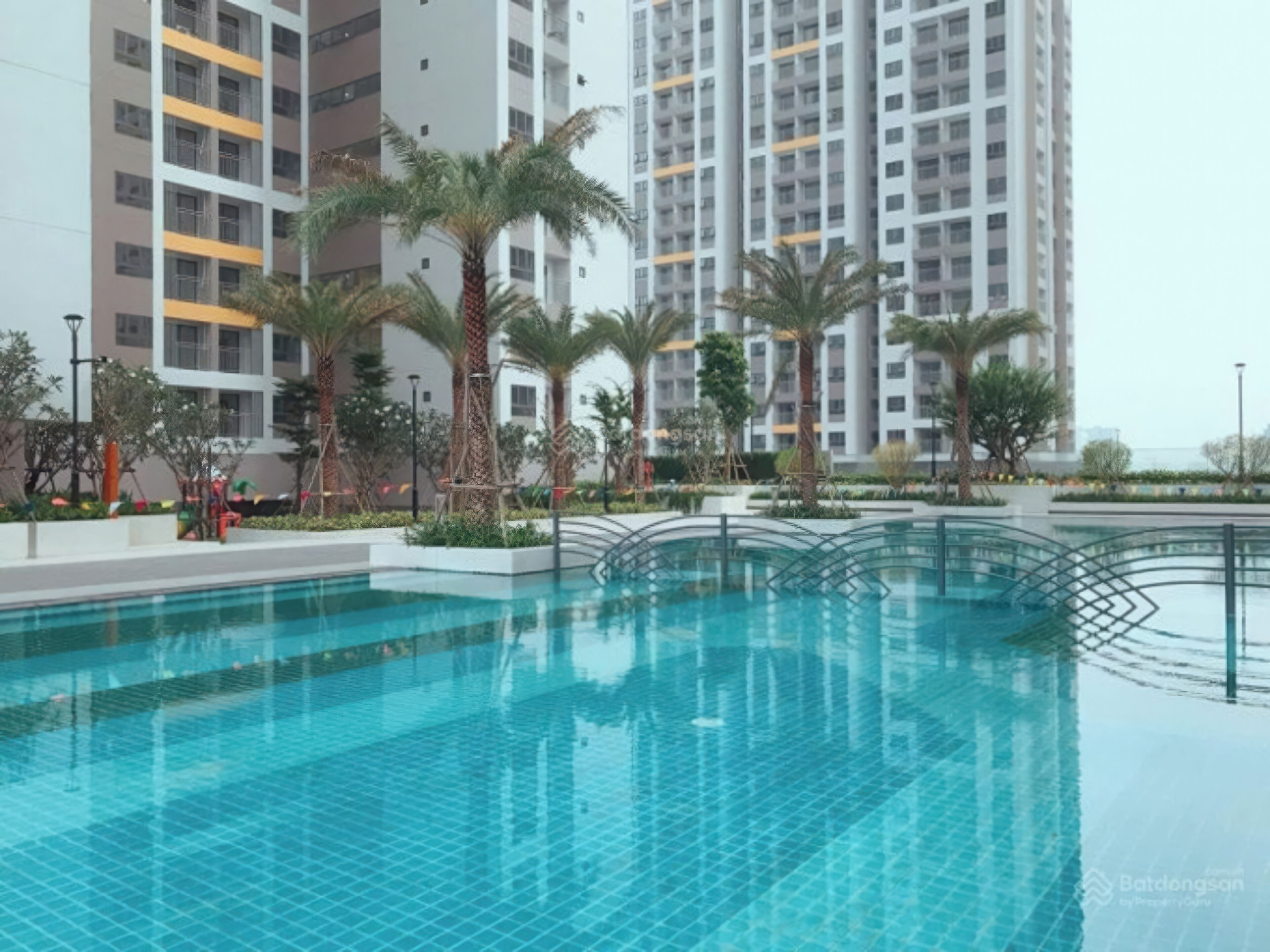 A swimming pool with a tiled deck, surrounded by palm trees and modern buildings in the background.