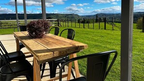 Une table en bois, entourée de chaises, placée sous une zone abritée, avec vue sur un champ verdoyant, une clôture et des collines lointaines.