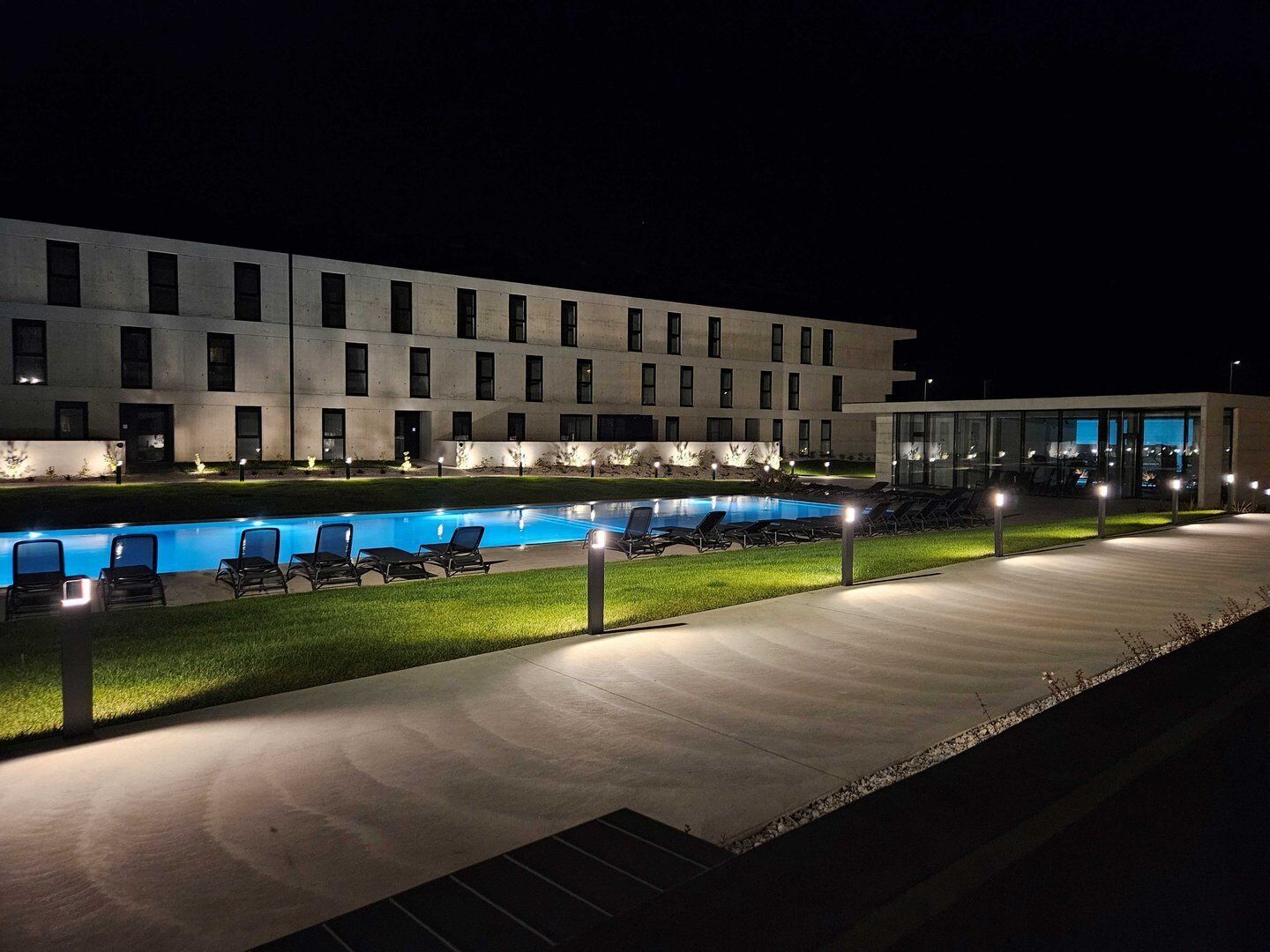 A modern hotel with a swimming pool, lounge chairs, and a well-lit walkway.