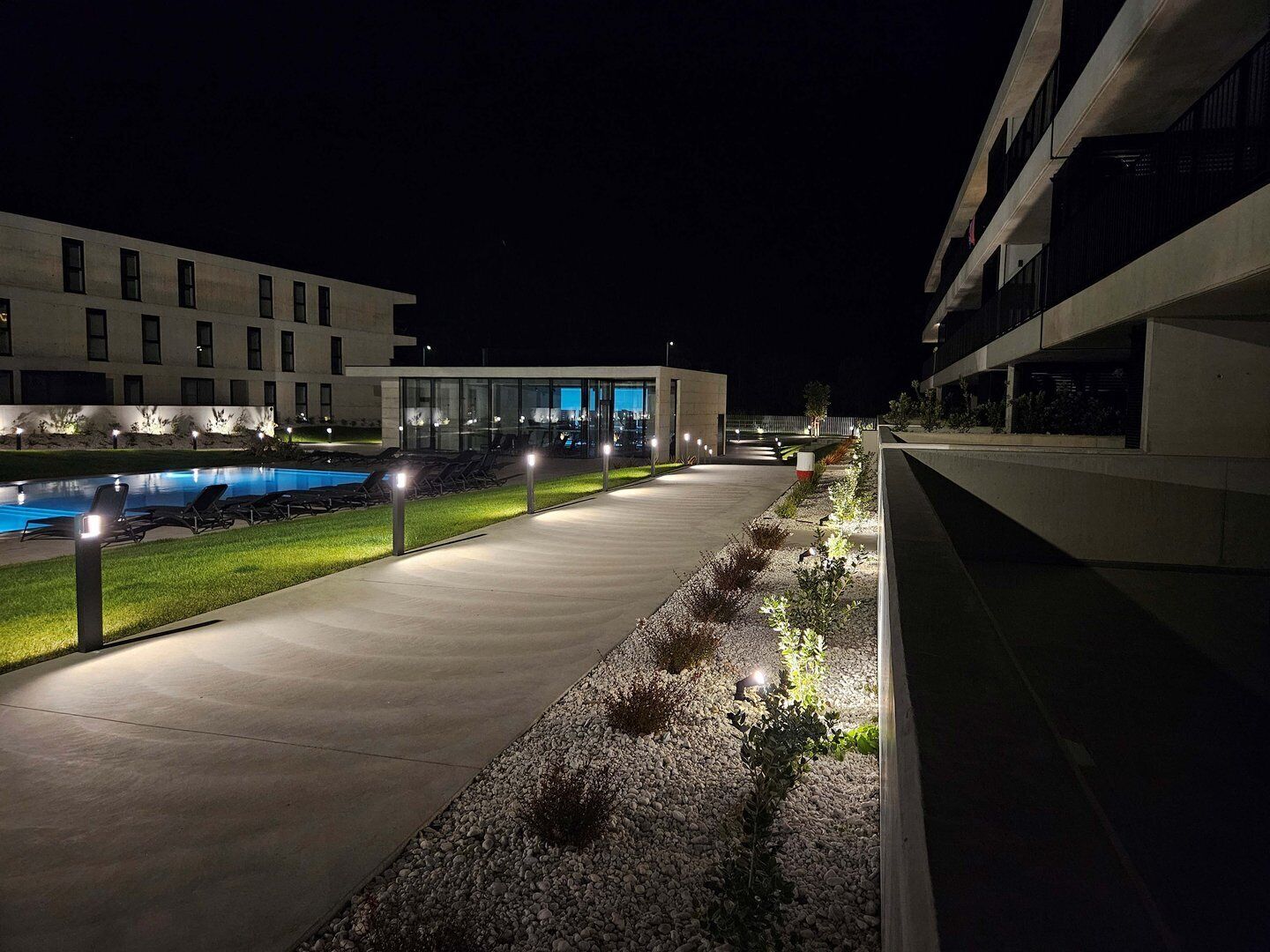 A modern outdoor pool area with a concrete pathway, lounge chairs, and a building with large windows in the background.