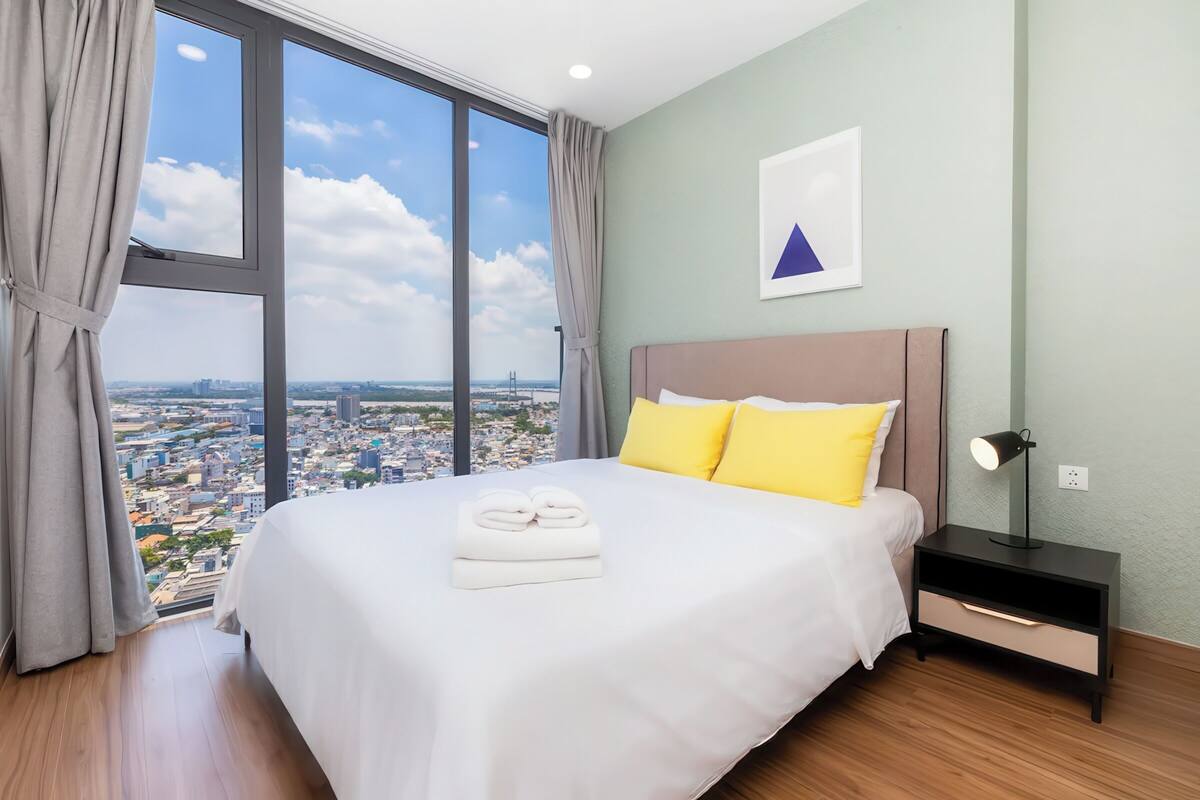 Une chambre d’hôtel avec un grand lit, une table de chevet, une lampe et une vue sur le paysage urbain, visible à travers une grande fenêtre.