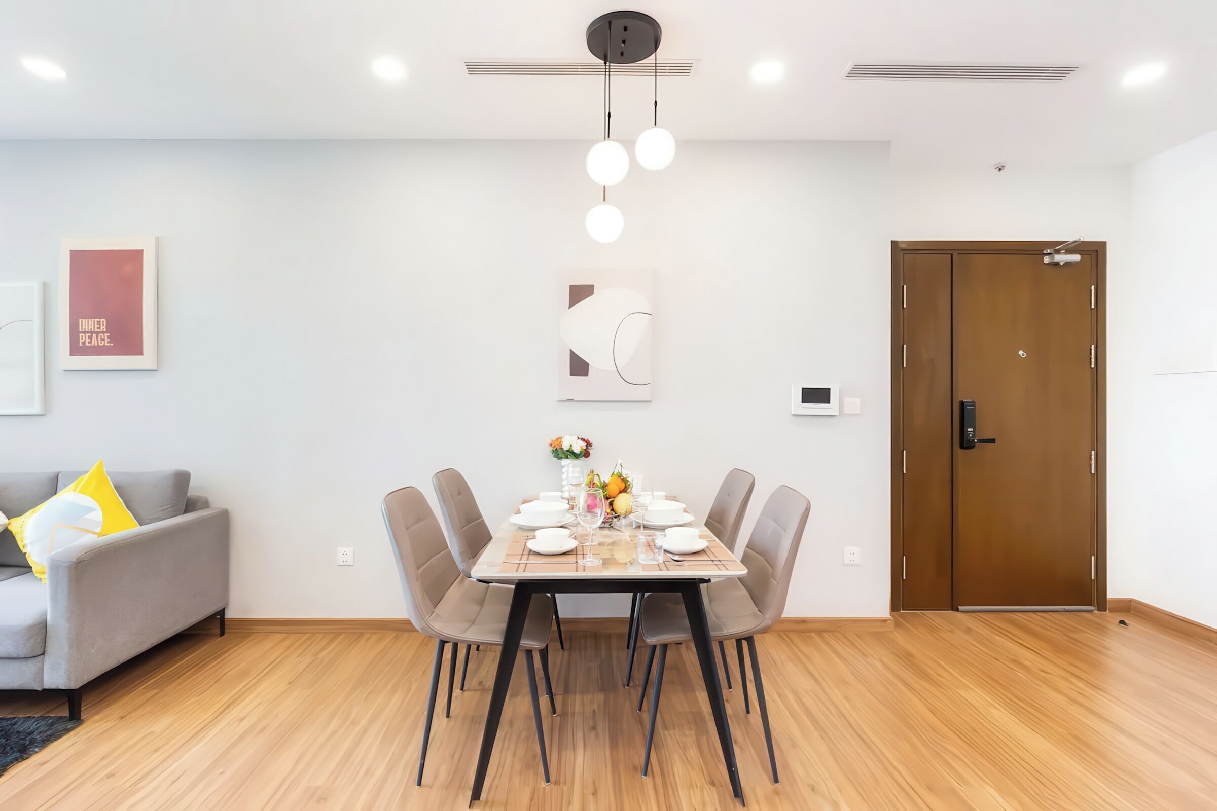 A modern dining area with a wooden table set for four, surrounded by grey chairs. A grey sofa with yellow pillows is against the wall, and a wooden door is visible to the right.