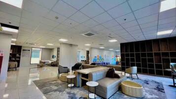 A spacious lobby with a modern design, featuring a seating area with sofas and round tables, a large window providing natural light, and a wall with a colorful mural.