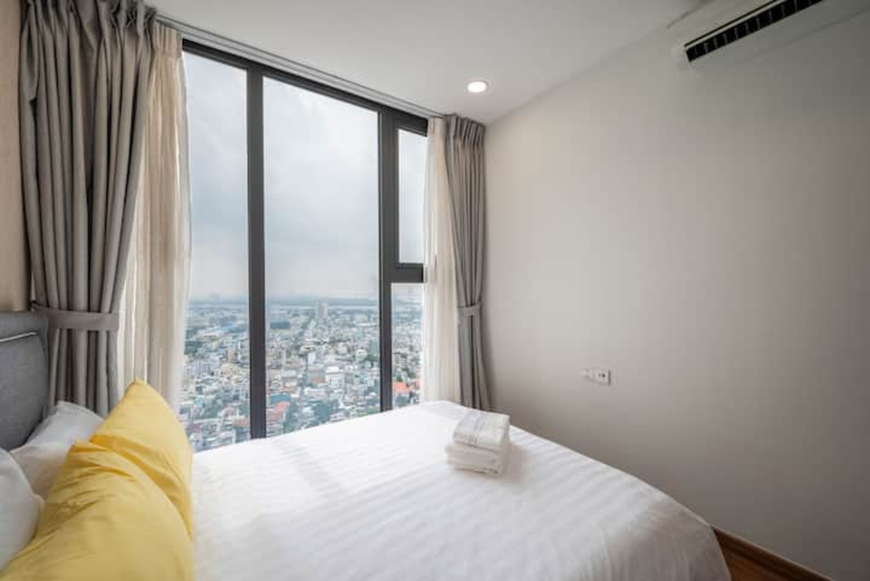 Habitación de hotel con un ventanal grande, una cama con ropa blanca y vistas a un paisaje urbano.