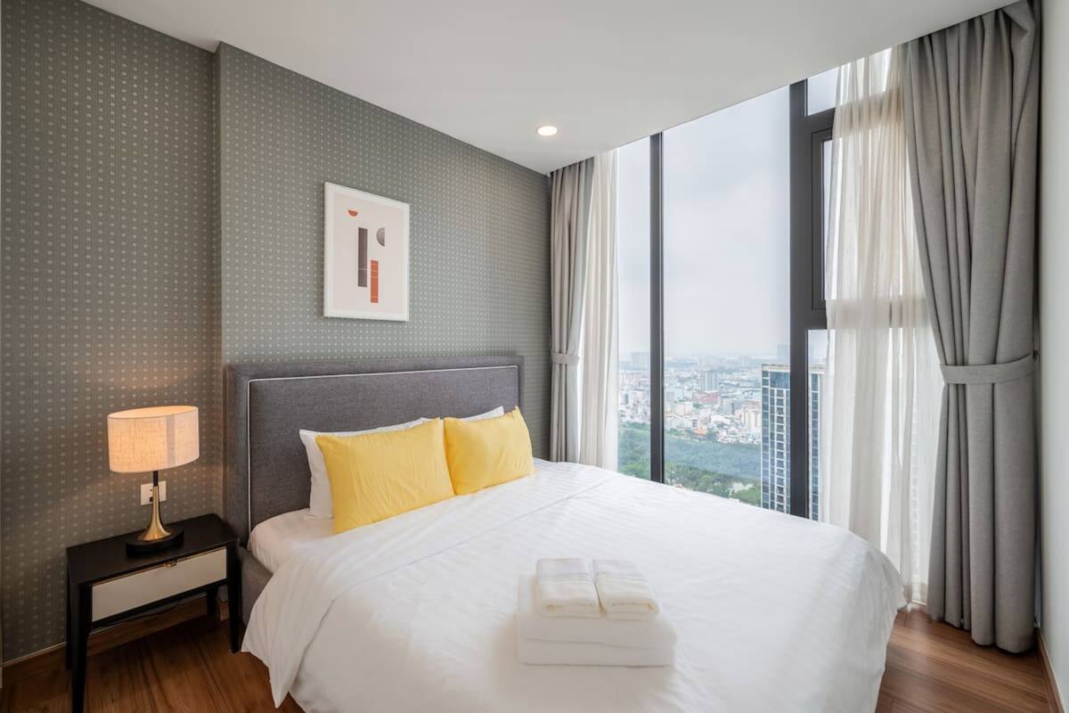 Une chambre d’hôtel avec un grand lit, une table de chevet, une lampe et une vue sur le paysage urbain, visible à travers de grandes fenêtres.