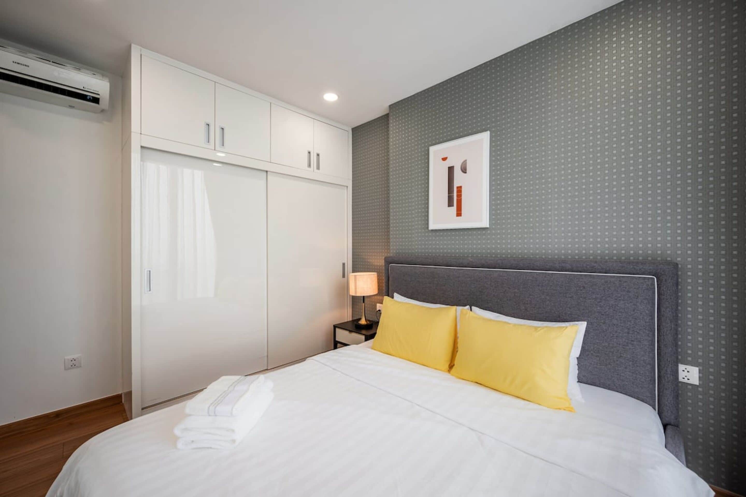 A hotel room with a bed, white curtains, a grey headboard, a wall-mounted artwork, and a white wardrobe.
