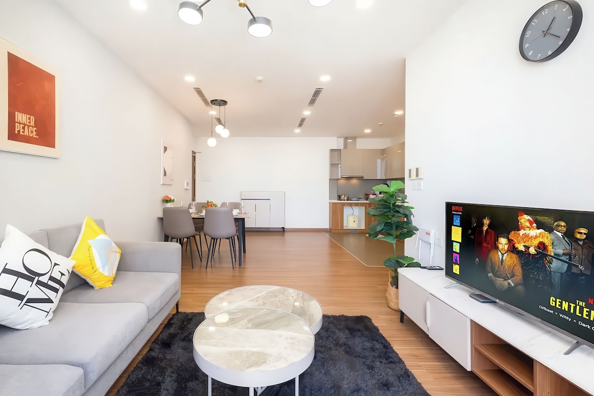 A modern living room with a grey sofa, a coffee table, and a flat-screen TV displaying 'The Gentlemen'. A dining area is visible in the background.
