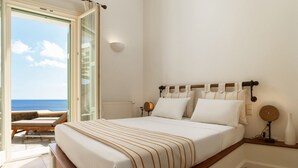 Un dormitorio con una cama grande, vista al mar, un balcón con mesa y sillas, y una lámpara de pared.