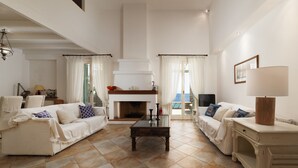 Amplia sala de estar con chimenea, muebles blancos y vistas al exterior.